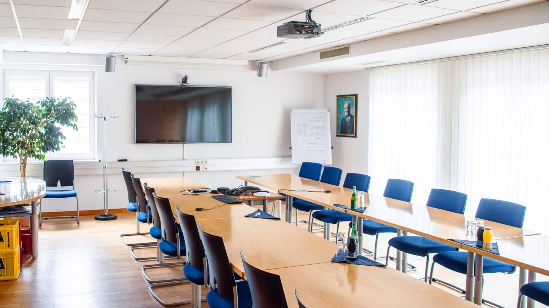 Heller Konferenzraum mit langen Holztischen, blauen Stühlen und moderner Präsentationstechnik.
