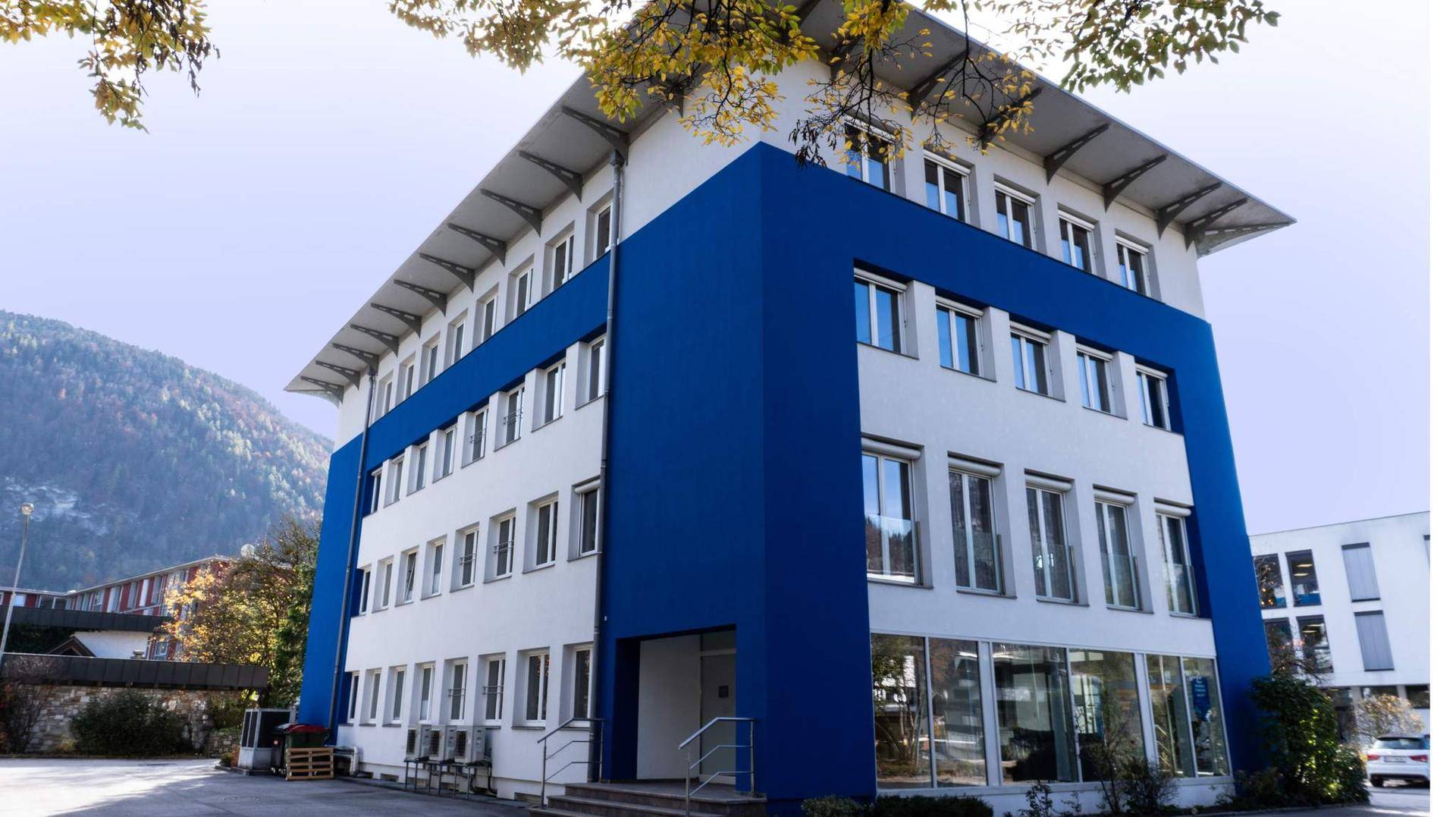 Seitenansicht des Bürogebäudes OFFICE 57 mit blauen und weißen Fassadenelementen vor grüner Bergkulisse.