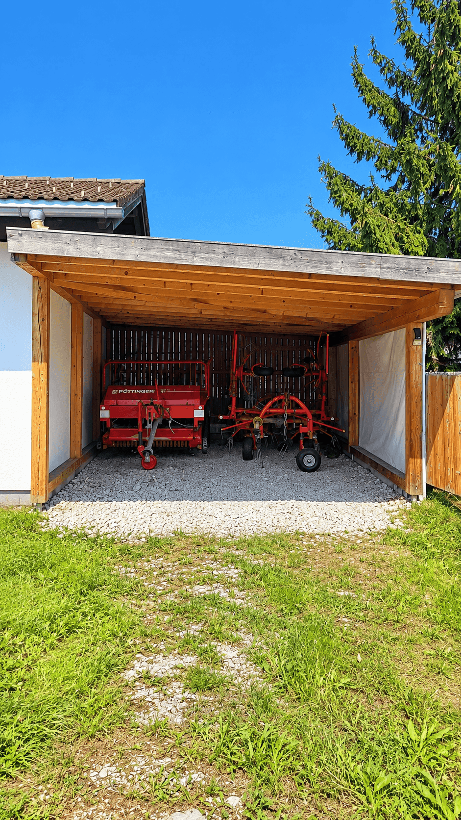 Offener Holzcarport mit landwirtschaftlichen Geräten auf Kiesboden unter blauem Himmel.