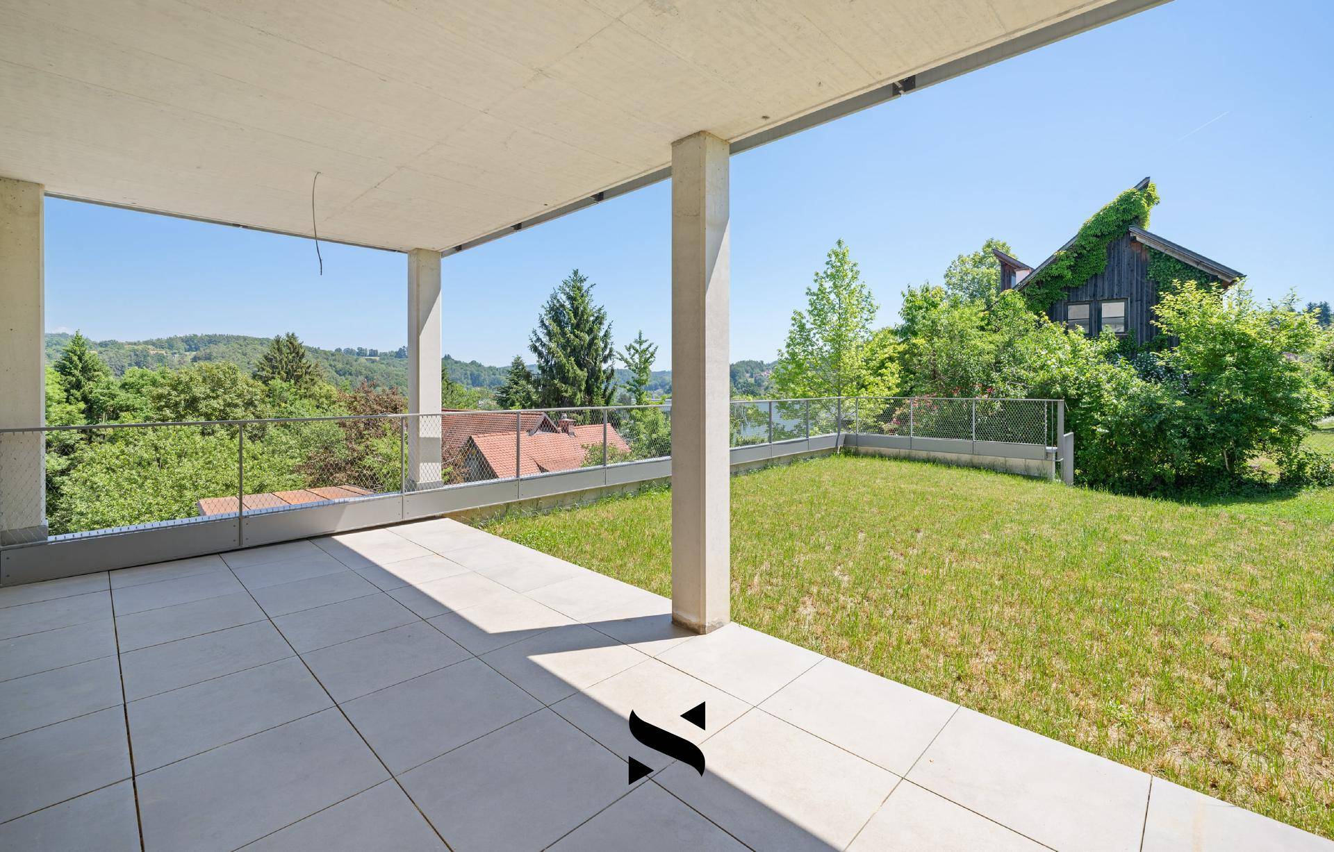 Überdachte Terrasse mit Fliesenboden und angrenzender Rasenfläche, umgeben von viel Grün.