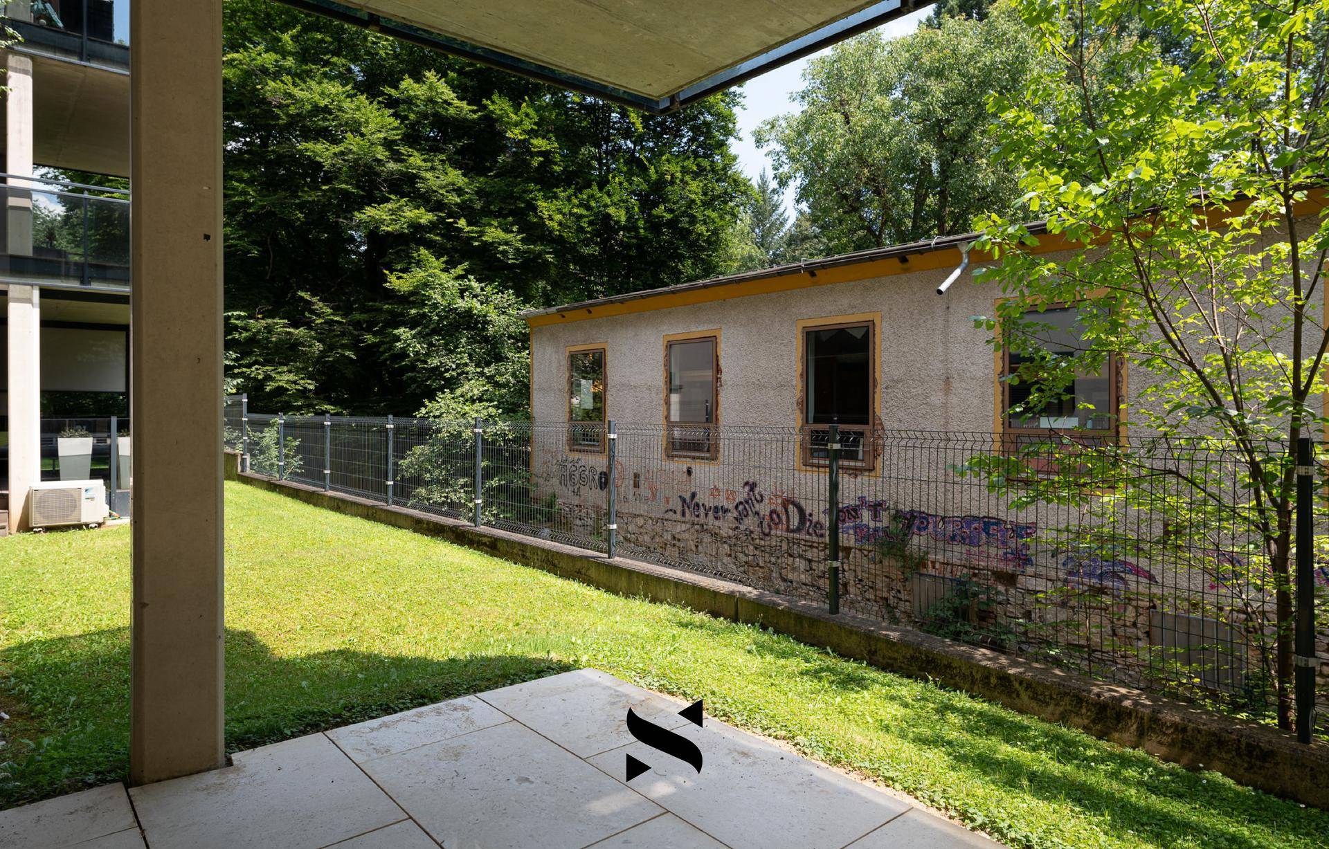 Grüner Gartenbereich mit Rasenfläche und Blick auf ein angrenzendes Gebäude mit Graffiti.