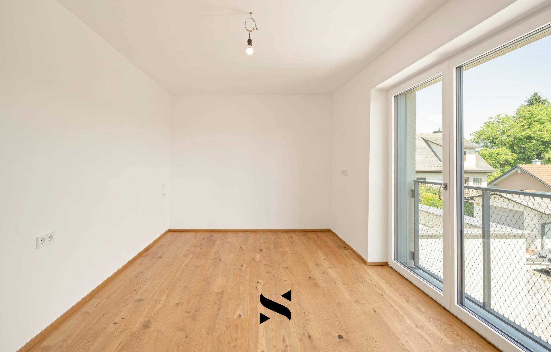 Leeres Zimmer mit Holzboden und Zugang zu einem französischen Balkon mit Blick auf die Umgebung.