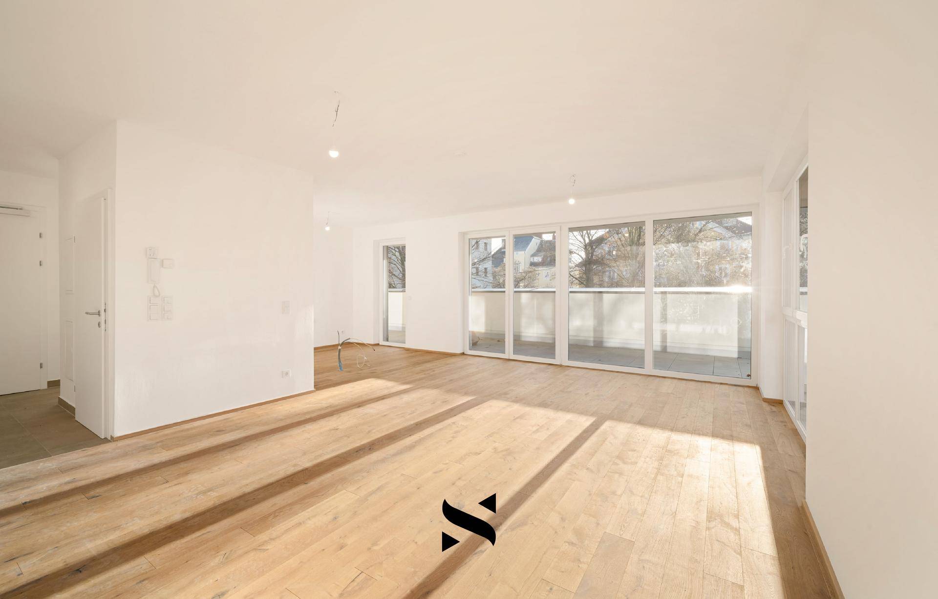Helles, leeres Wohnzimmer mit Holzboden und großen Fenstern zum Balkon.