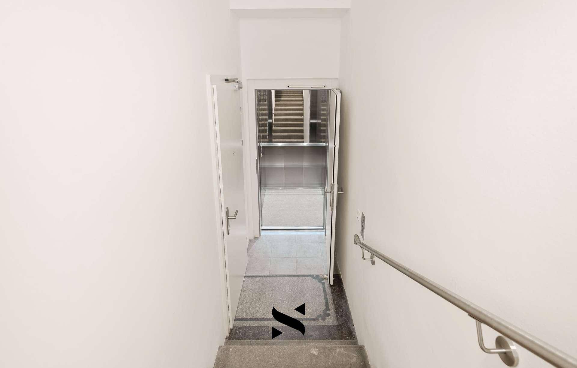 Heller Treppenaufgang mit Handlauf und Zugang zu einem modernen Aufzug.