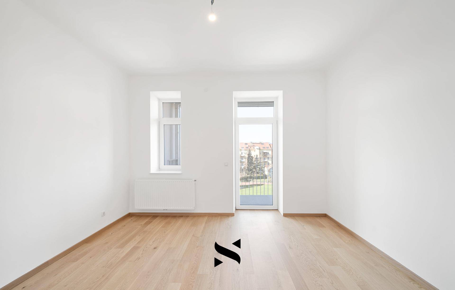 Leeres Zimmer mit Parkettboden, weißen Wänden und Zugang zu einem französischen Balkon.