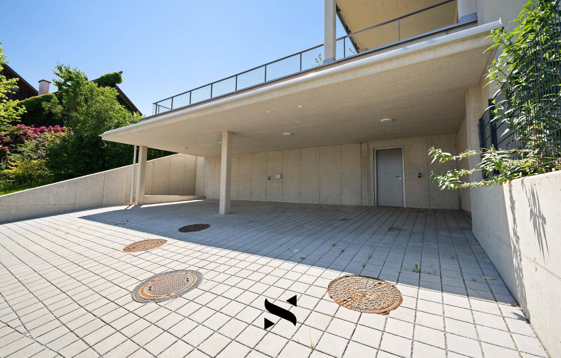 Überdachter Parkplatz oder Carport mit gepflastertem Boden vor dem Gebäude.