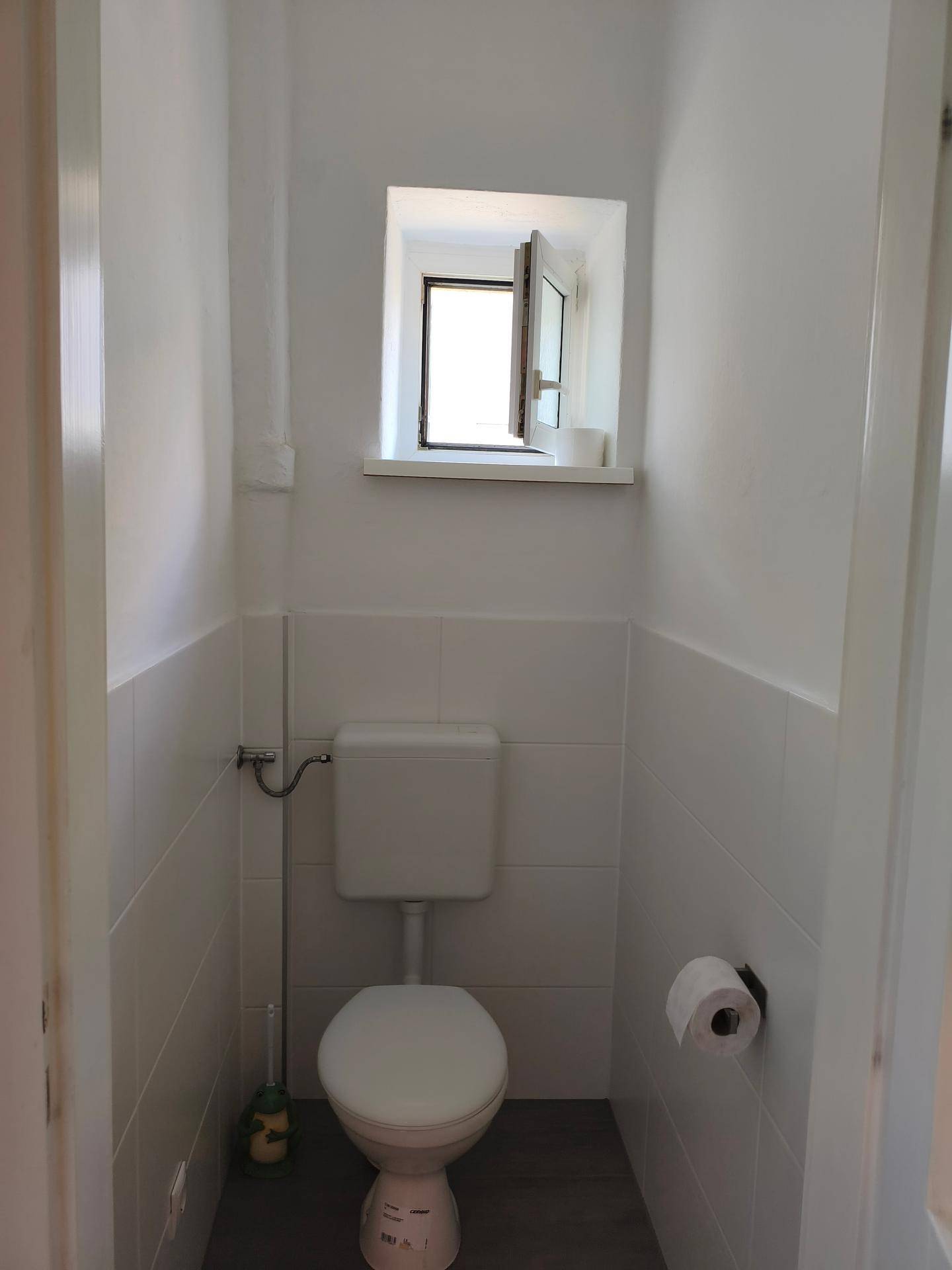 Separates WC mit Fenster, weiß gefliesten Wänden und einfacher Toilette.