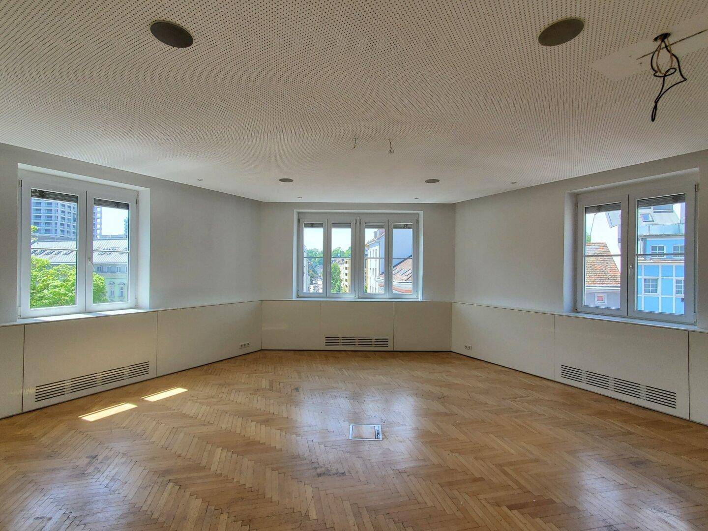 Ein geräumiger, leerer Büroraum mit Fischgrätparkett, hohen Decken und großen Fenstern, die viel Licht hereinlassen.