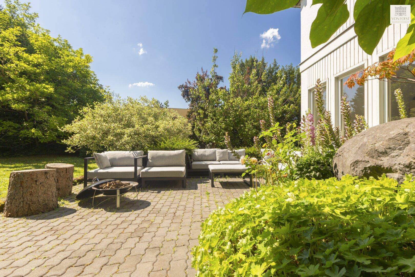 Geräumige Terrasse mit moderner Lounge-Möblierung und Feuerstelle, umgeben von üppigem Grün.