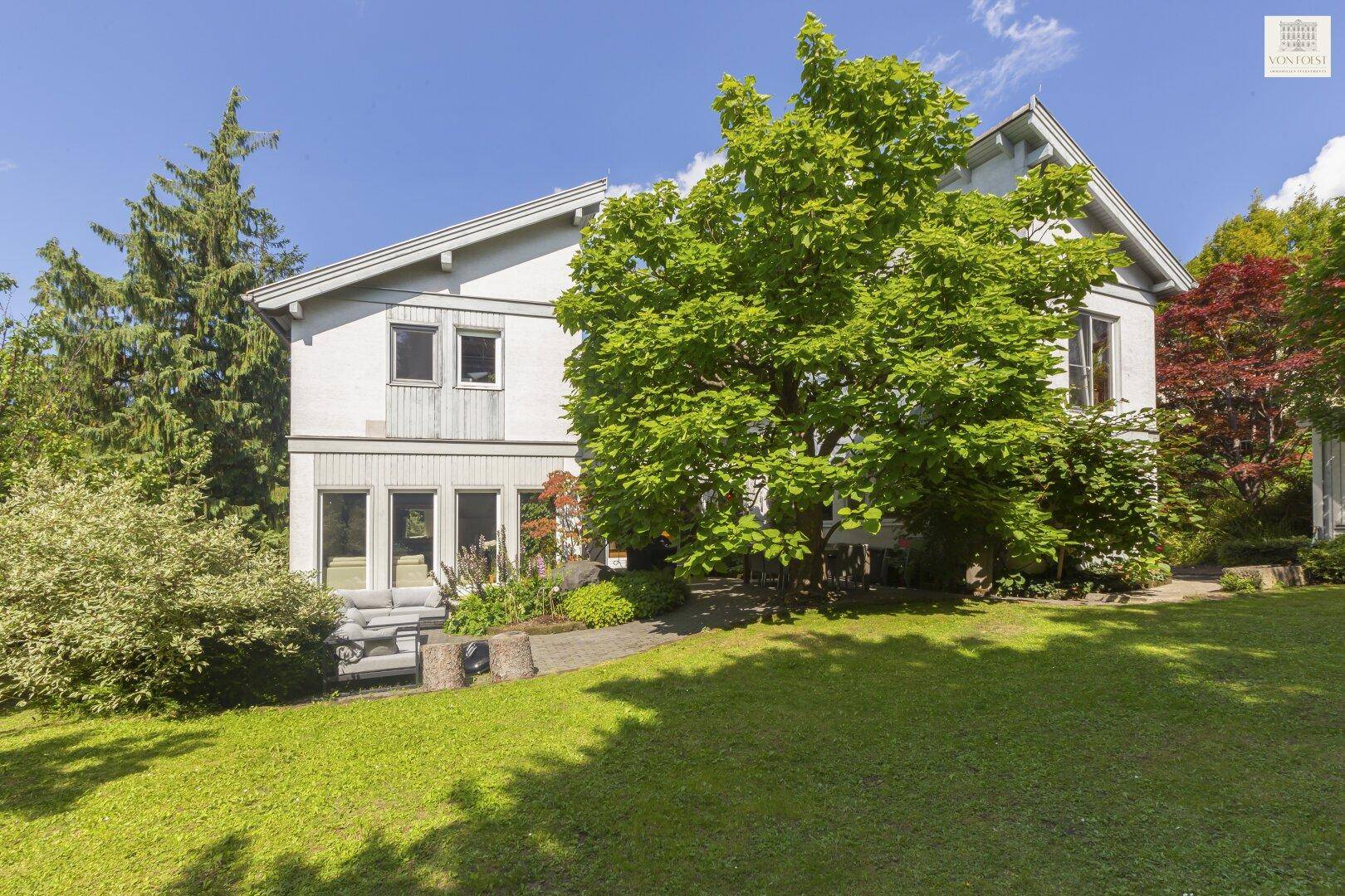 Ansicht des Hauses mit gepflegtem Rasen und hohen Bäumen unter blauem Himmel.