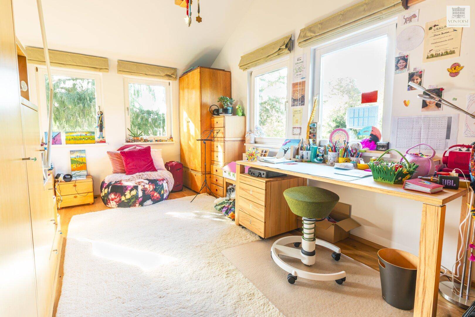 Helles Kinderzimmer mit Schreibtisch, Teppichboden und großen Fenstern mit Grünblick.