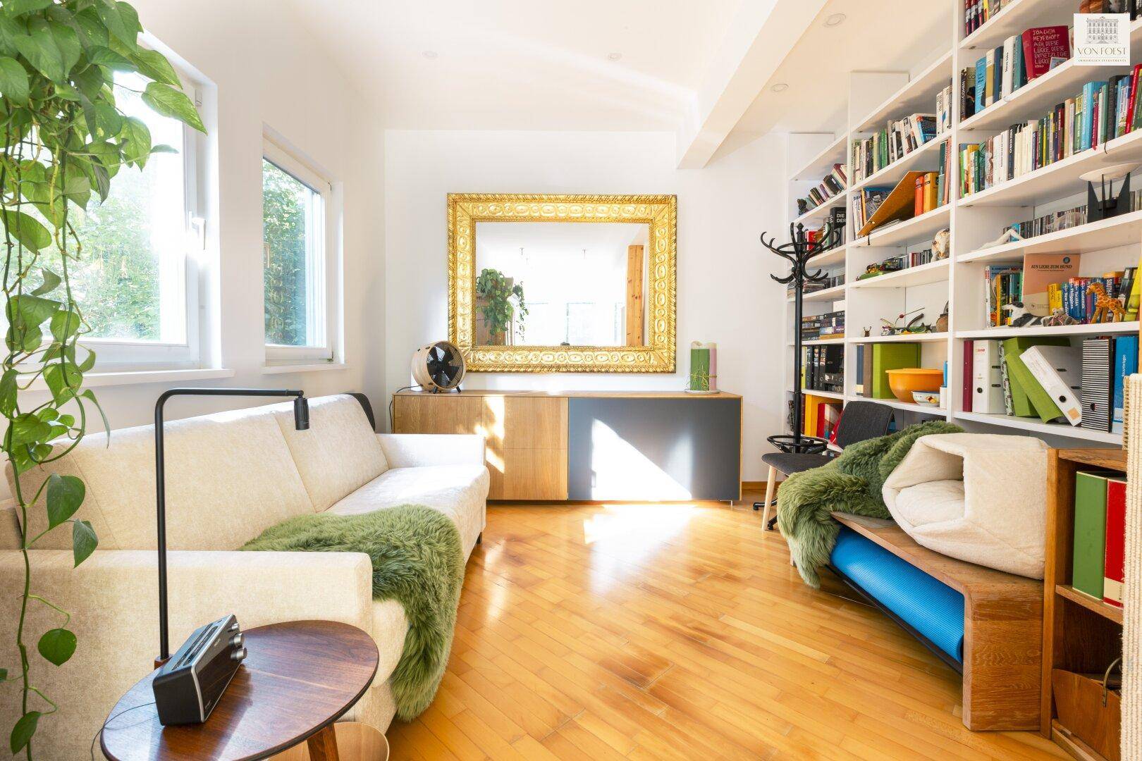 Helles Wohnzimmer mit Bücherregal, beigem Sofa und großem Spiegel, Holzboden.