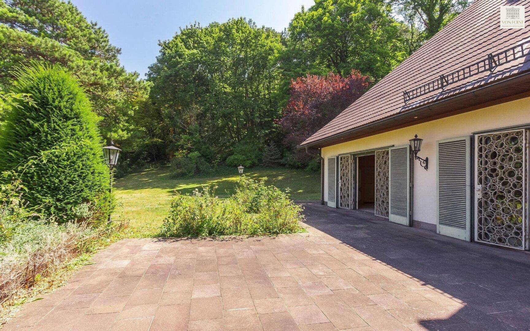 Geräumige gepflasterte Terrasse am Haus, umgeben von Grünflächen und hohen Bäumen.