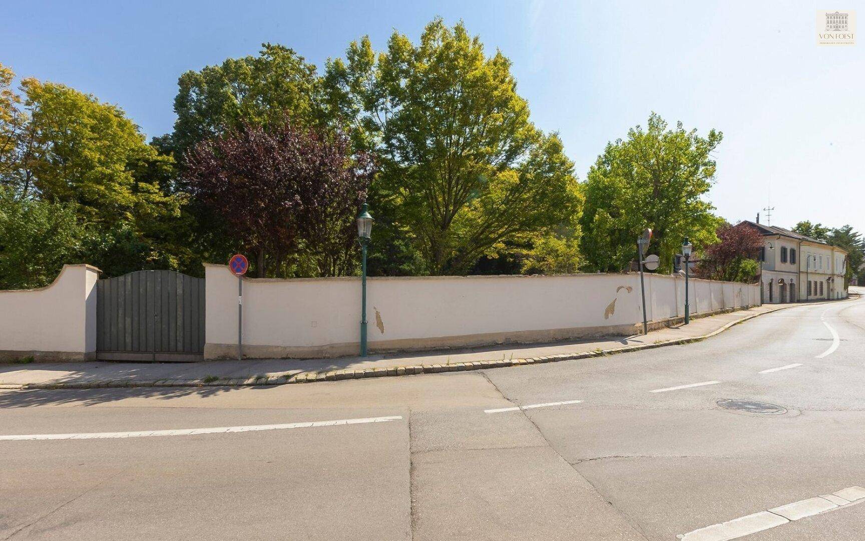Lange Grundstücksmauer mit Holztor und Blick auf die Straße sowie weitere Gebäude in der Ferne.