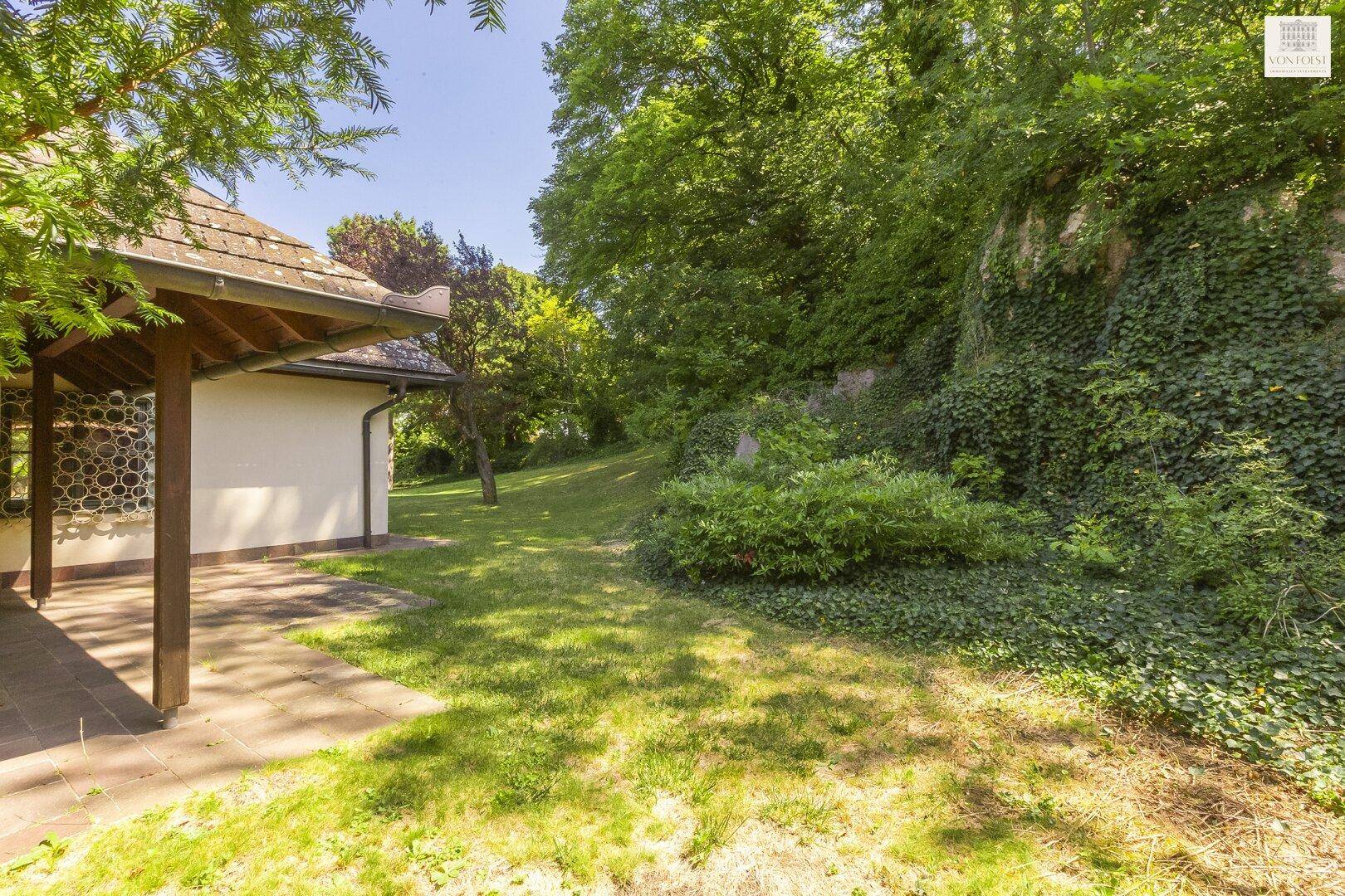 Überdachter Terrassenbereich am Haus, angrenzend an einen grünen Hang mit üppiger Vegetation.