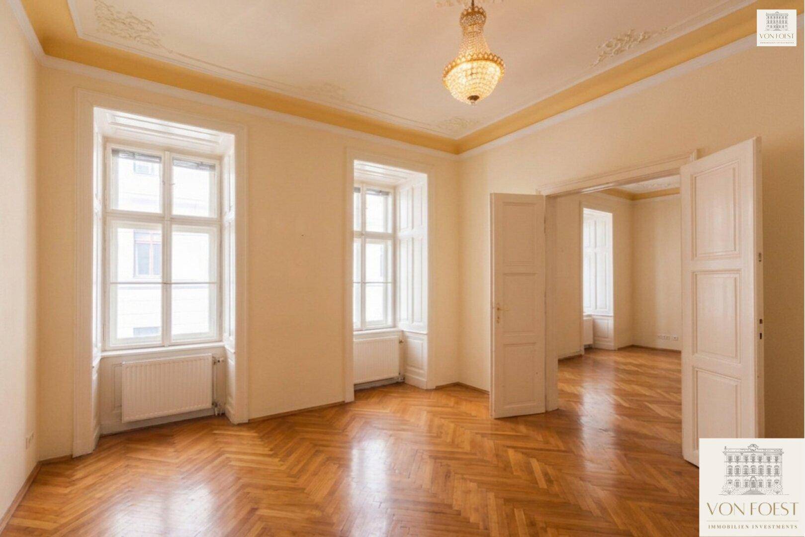 Helles Zimmer mit Fischgrätparkett, hohen Fenstern und klassischer Stuckverzierung an der Decke.