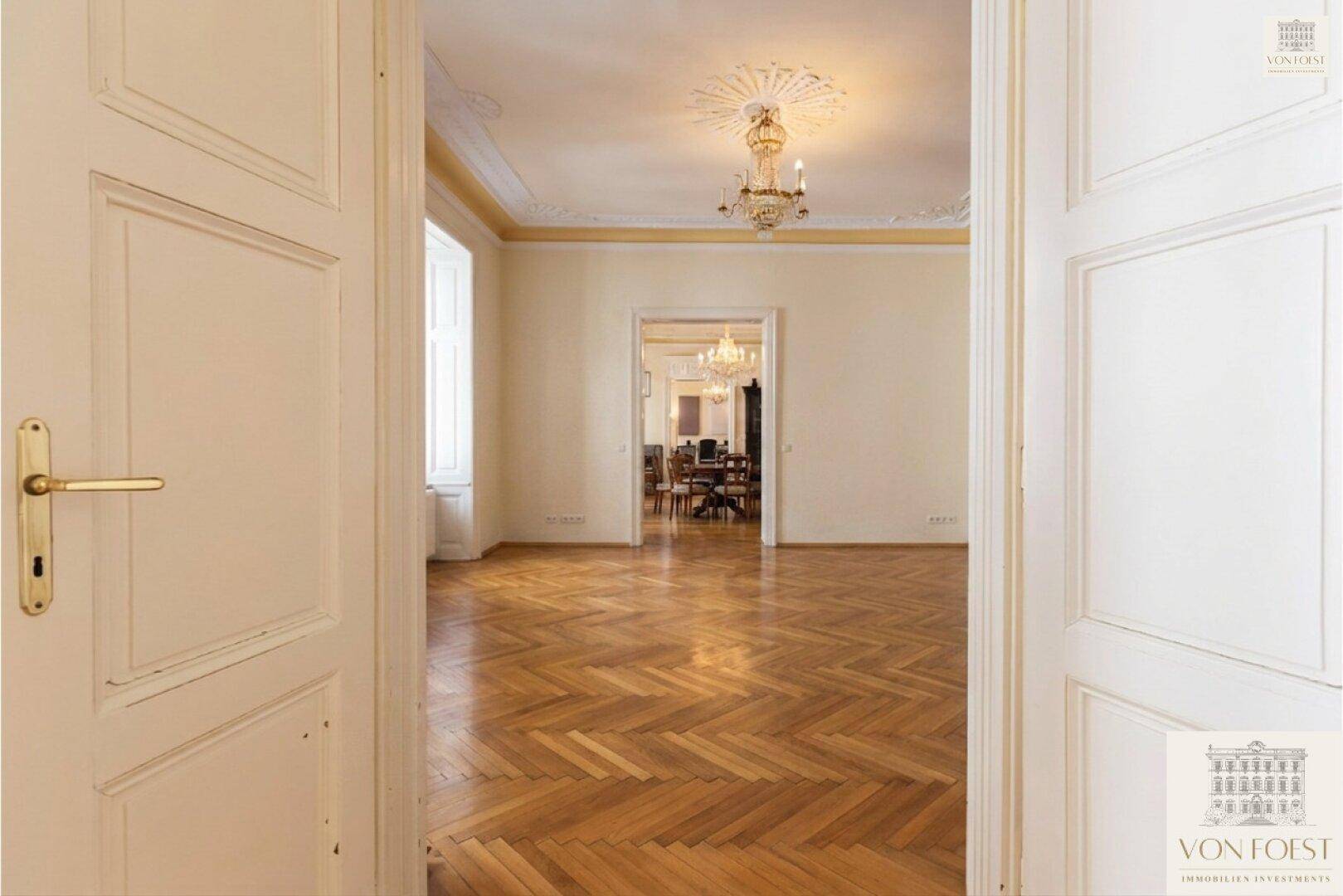 Blick durch eine offene Tür in ein weiteres Zimmer mit Fischgrätparkett und Kronleuchter.
