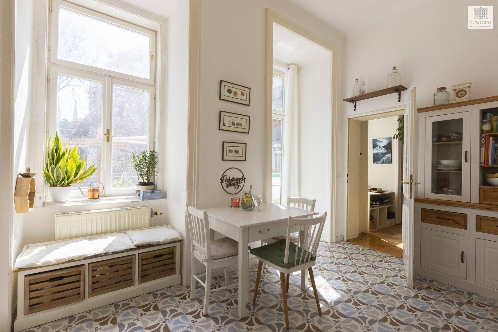 Helle Wohnküche mit Esstisch, gemusterten Fliesen und Fensterbank mit Pflanzen.
