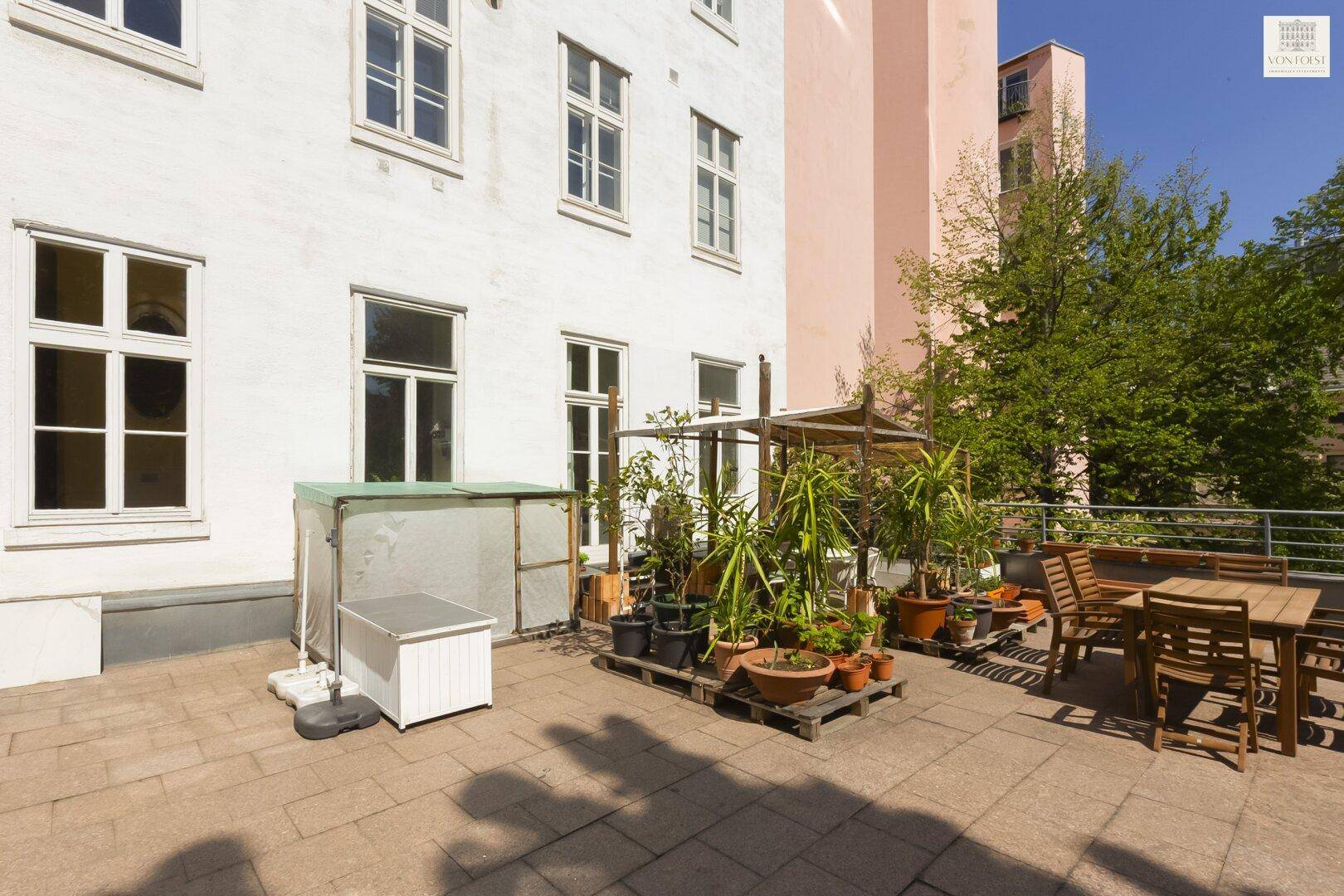 Große gepflasterte Terrasse mit vielen Pflanzen und Gartenmöbeln am Gebäude.