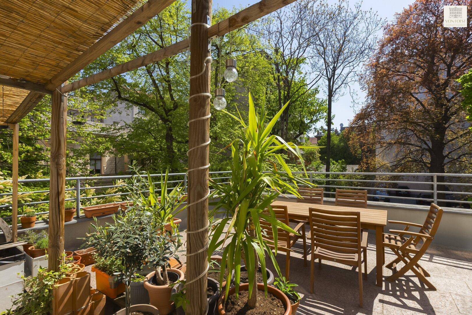 Sonnige Terrasse mit Holzmöbeln, vielen Pflanzen und Bambusdach.