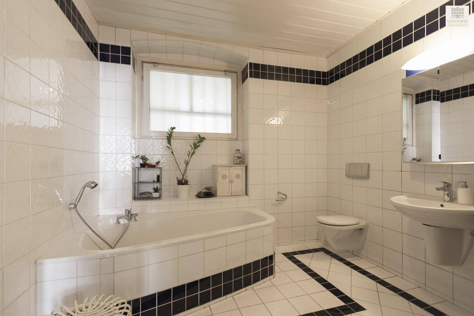 Badezimmer mit Badewanne, WC und weißen Fliesen mit schwarzen Akzenten.
