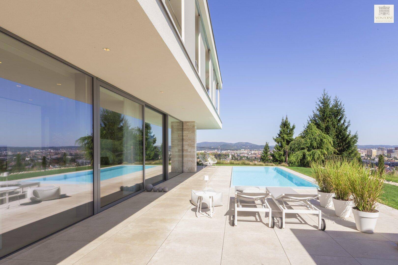 Entspannungsbereich am Pool mit Sonnenliegen und weitem Blick über die Landschaft und Stadt.