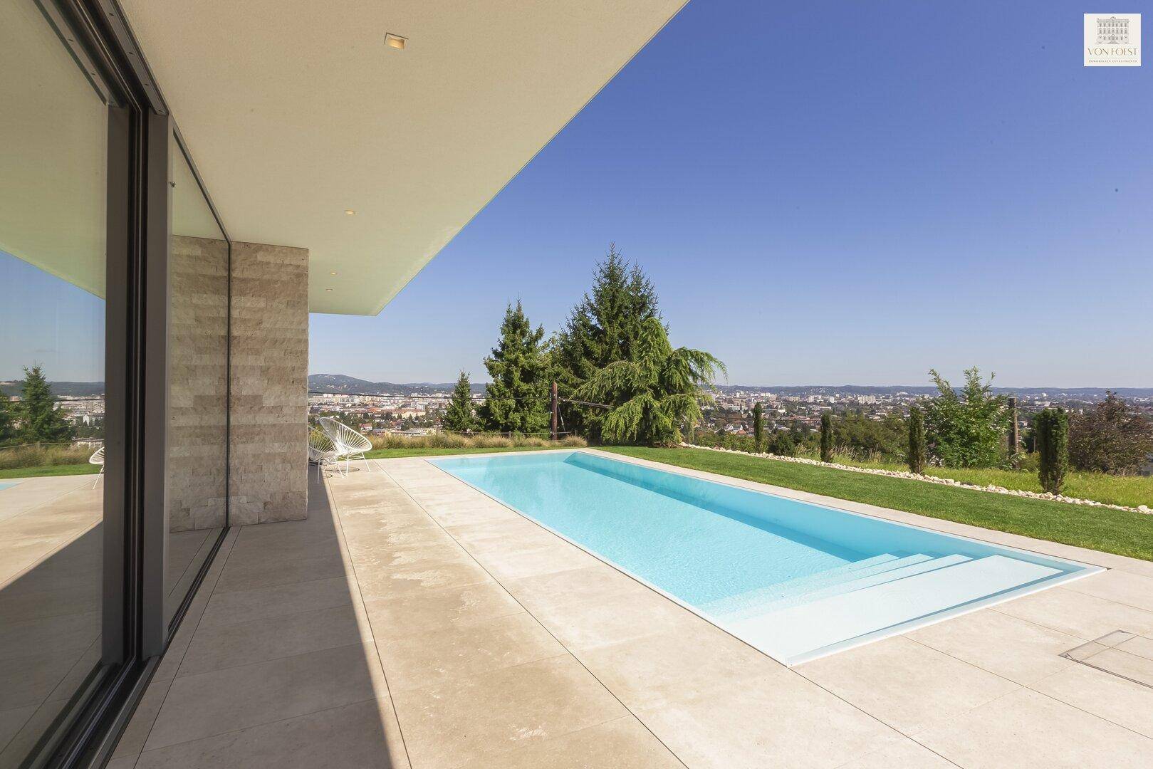 Langer, rechteckiger Pool mit Stufen, umgeben von einer hellen Terrasse und Grünflächen.