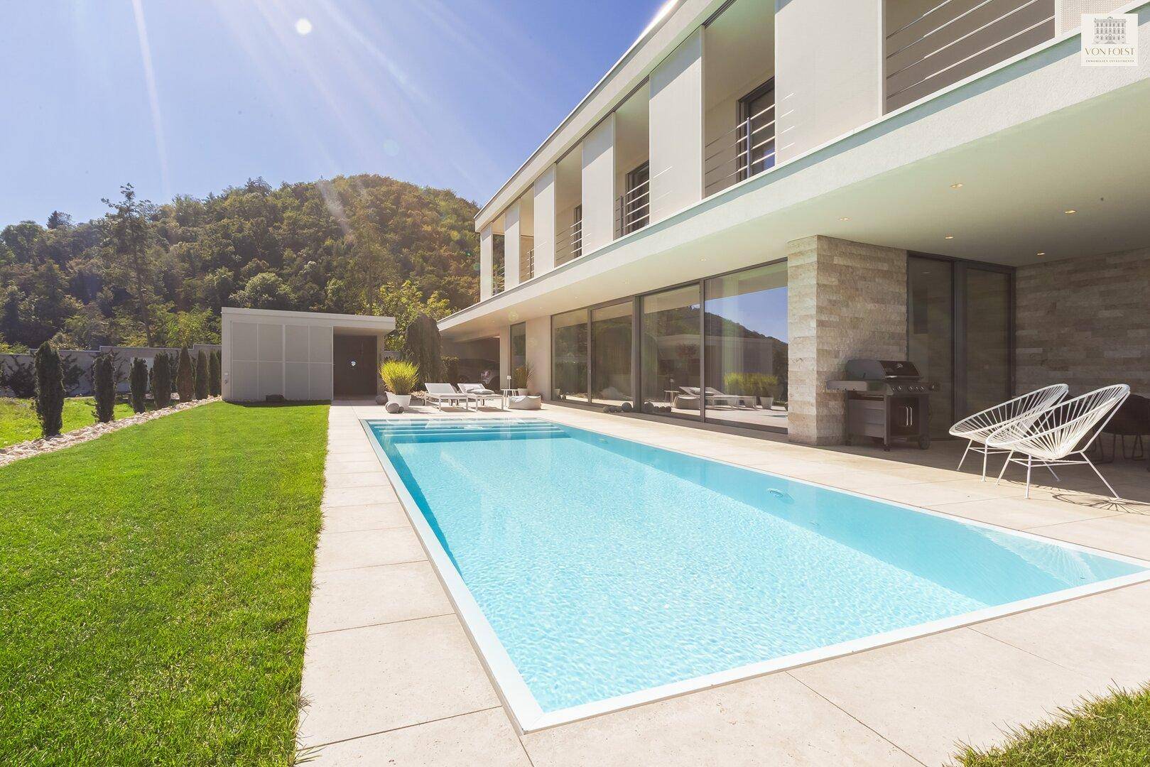 Modernes Haus mit großem Swimmingpool, weitläufigem Rasen und Bergblick unter strahlendem Himmel.
