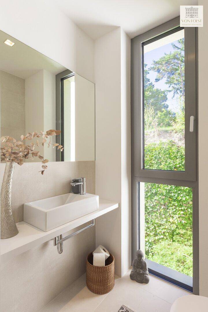 Modernes Badezimmer mit rechteckigem Waschbecken, großem Spiegel und Fenster mit Blick ins Grüne.