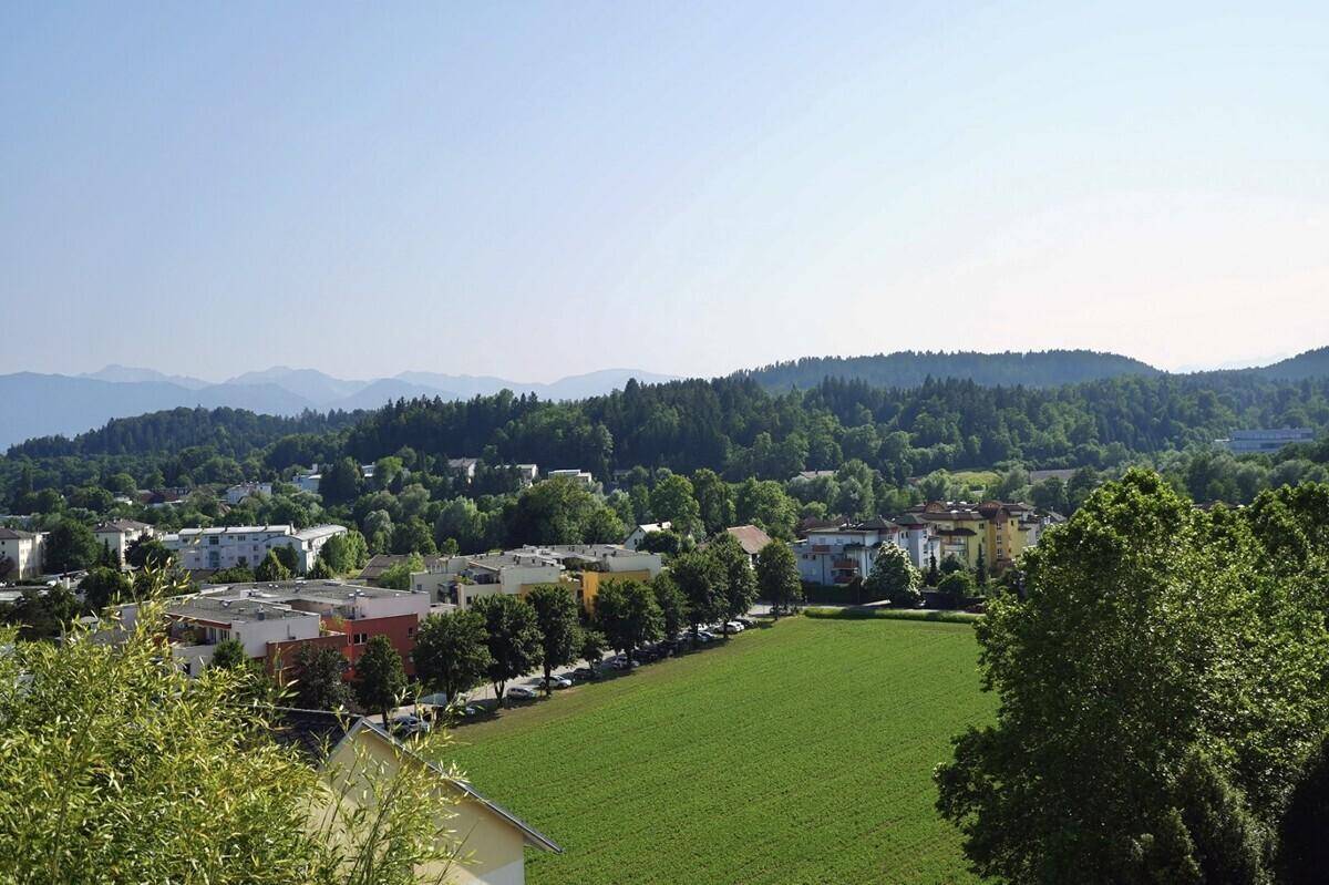 Weitläufiger Panoramablick über eine grüne Landschaft mit Siedlungen und bewaldeten Bergen im Hintergrund.