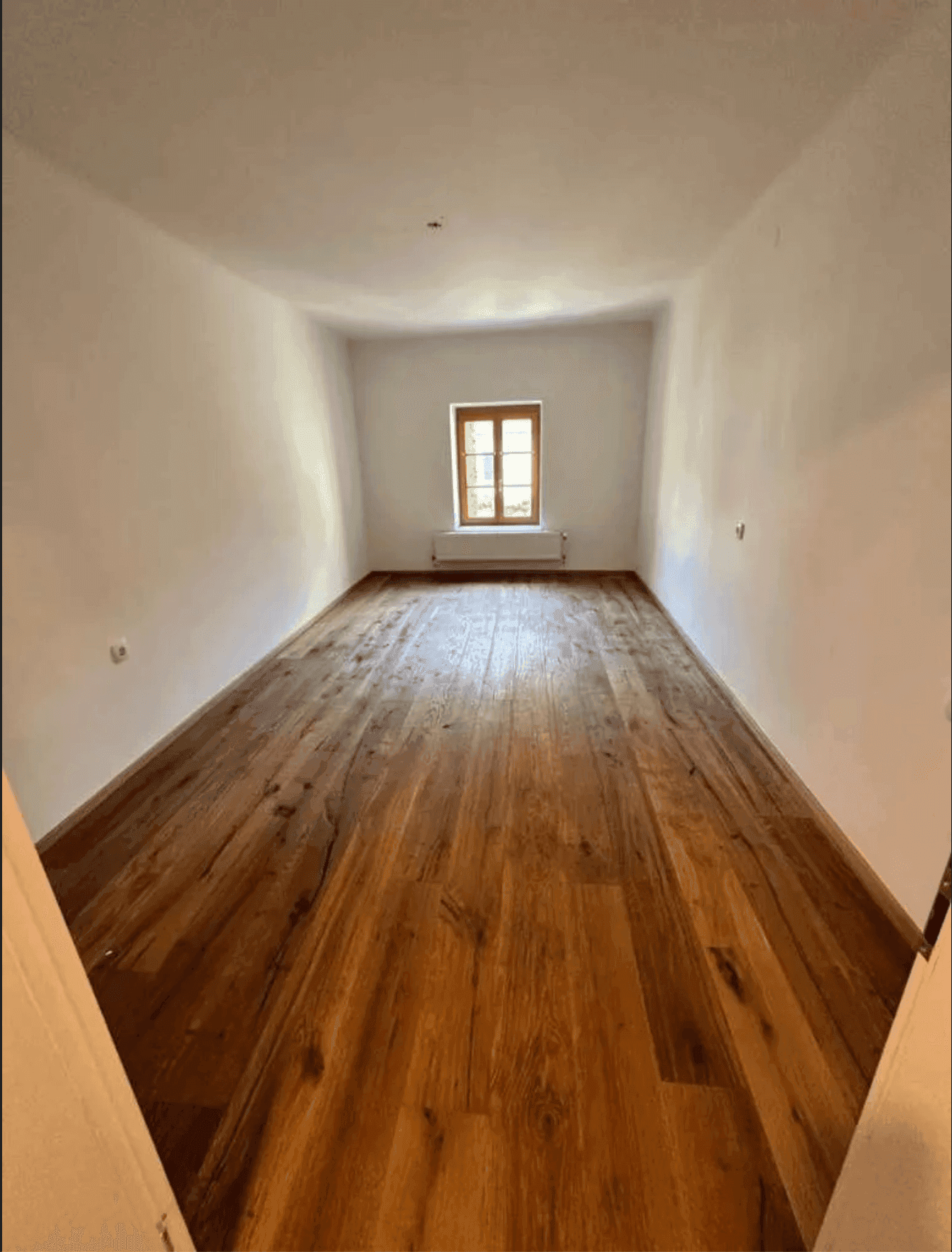 Langes, schmales Zimmer mit Holzboden und einem Fenster, ideal für ein Schlafzimmer oder Büro.