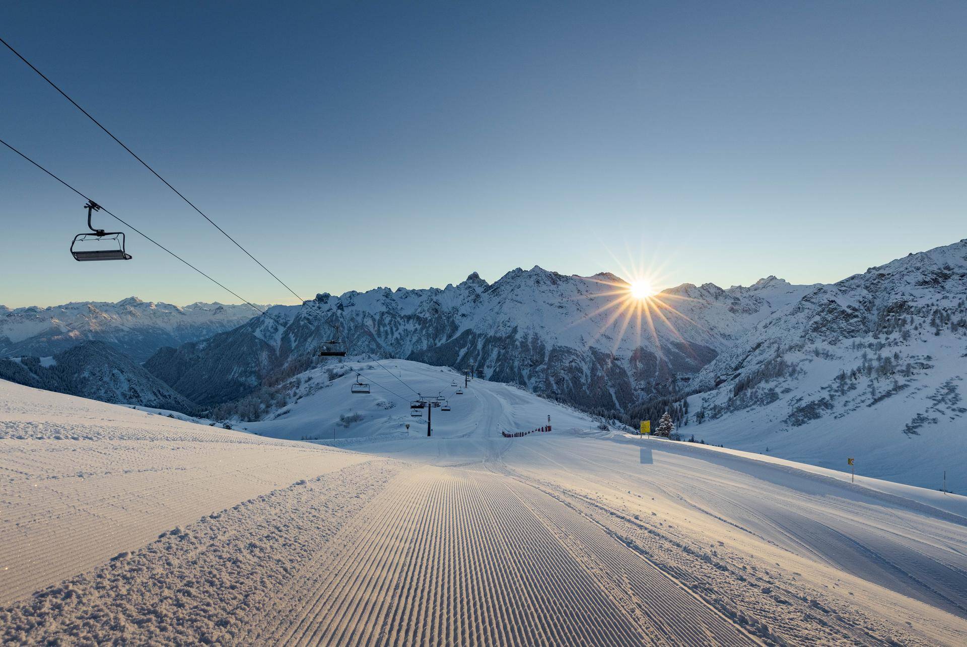Sonnenuntergang über einer präparierten Skipiste mit Skilift und majestätischen Bergen im Hintergrund.