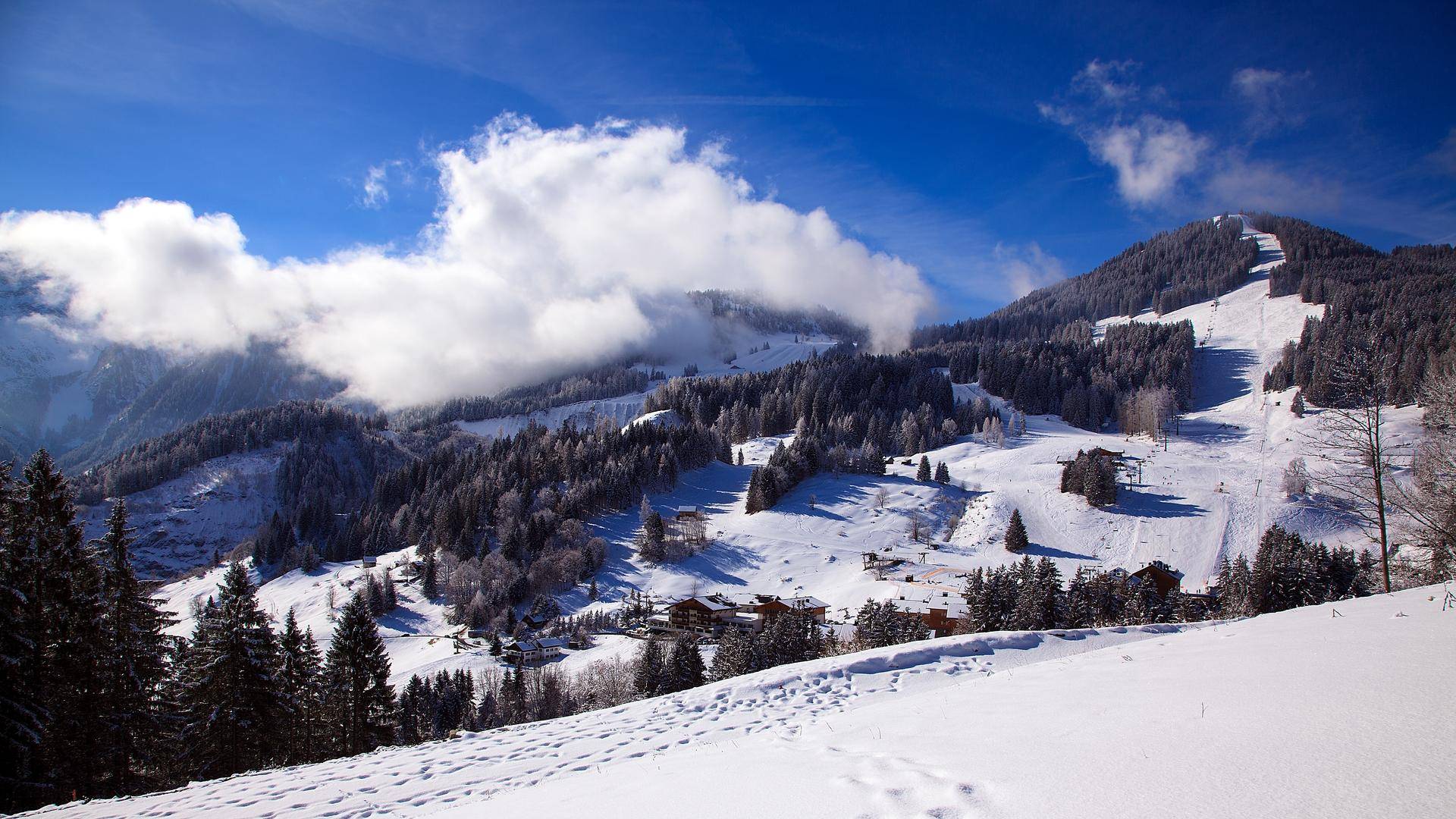 Winterliche Berglandschaft mit Wolken, verschneiten Wäldern und einer Skipiste in den Alpen.