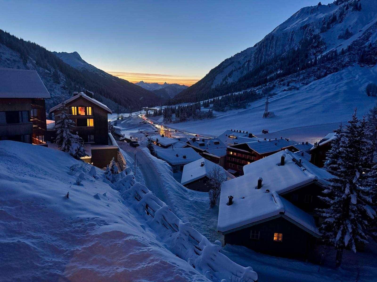 Verschneites Dorf in einem Bergtal bei Sonnenuntergang mit beleuchteten Häusern.