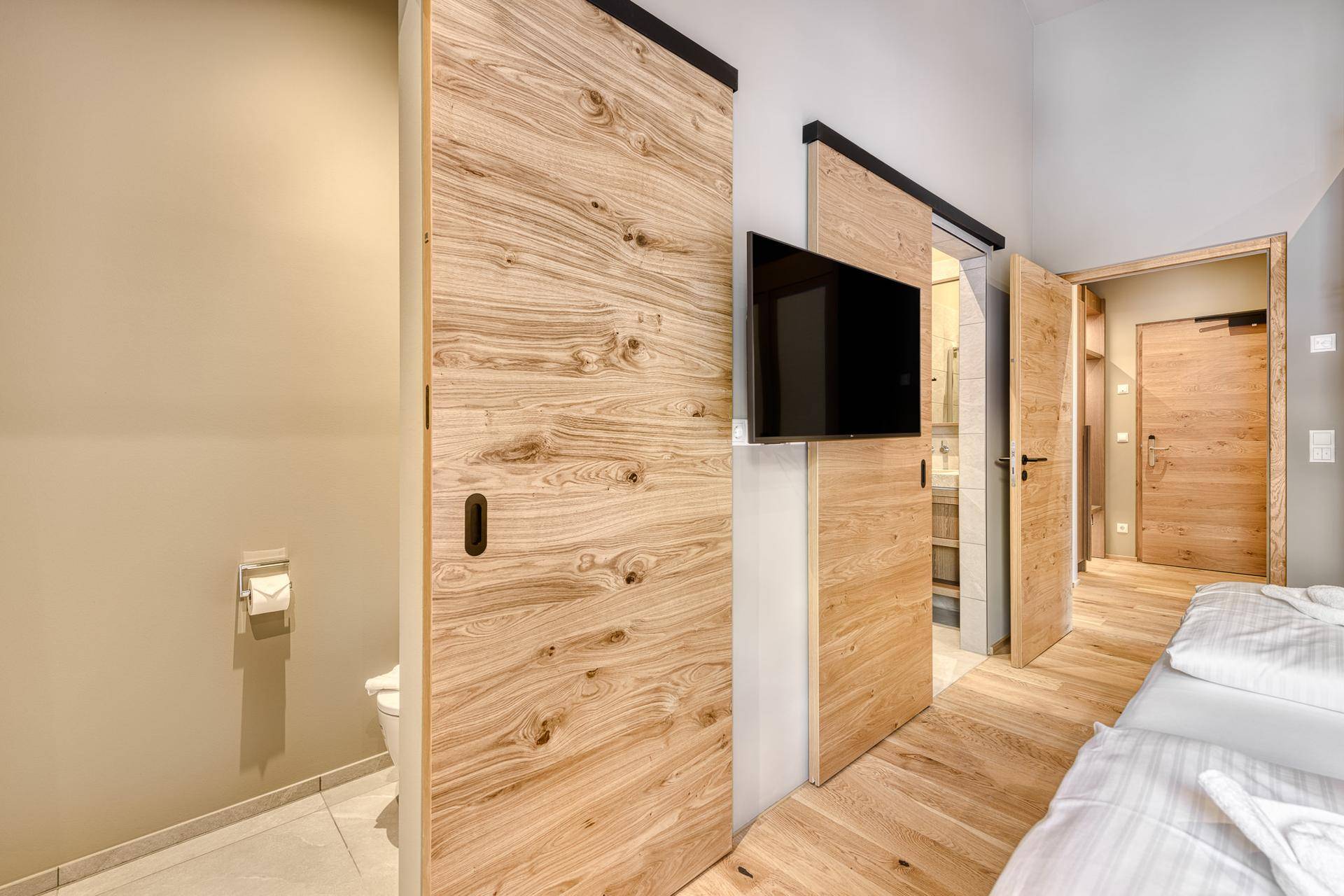 Schlafzimmer mit Holzboden, Wandfernseher und integriertem WC-Bereich hinter Schiebetüren.