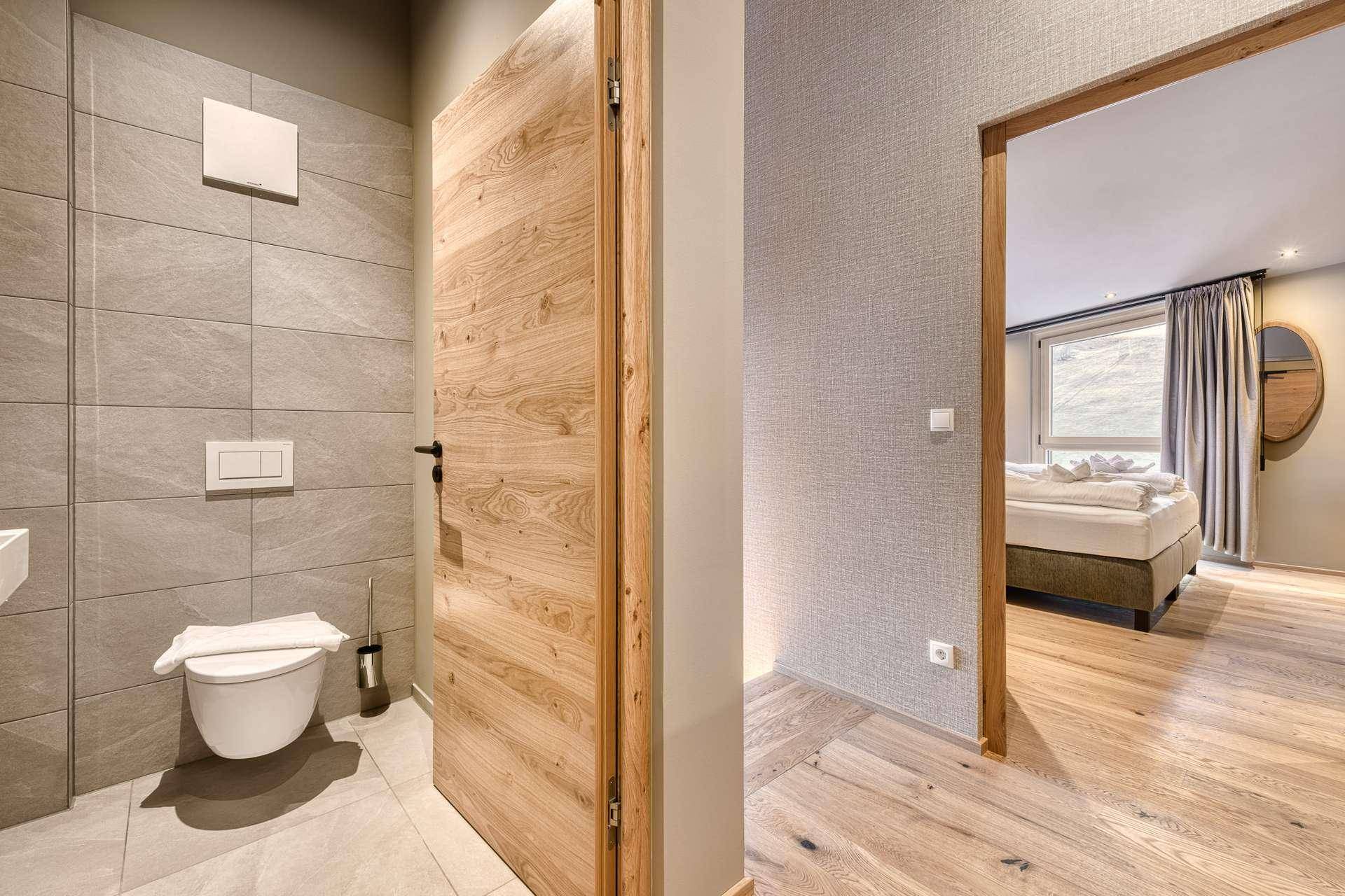Modernes WC mit Wand-WC und Fliesenboden, mit Blick in ein angrenzendes Schlafzimmer.