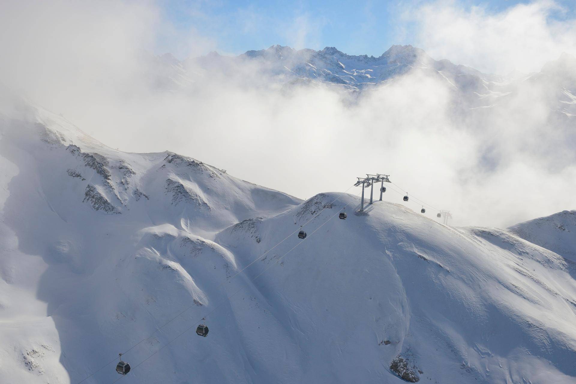 Verschneite Berglandschaft mit Skiliften und Gondeln, umgeben von Wolken und Gipfeln.