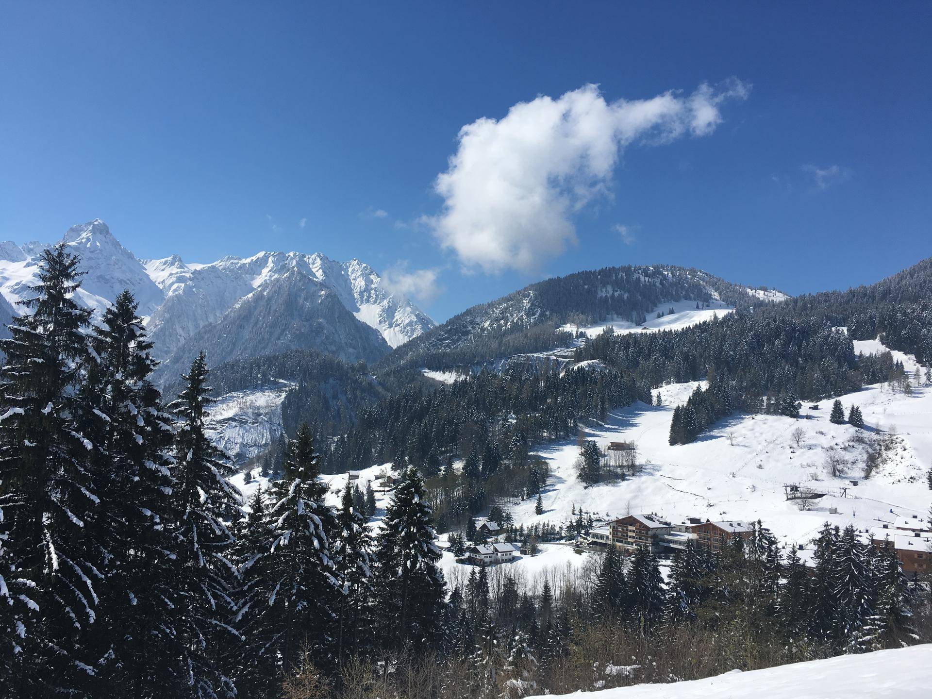 Winterliche Berglandschaft mit verschneiten Bäumen, einer Skipiste und vereinzelten Gebäuden unter blauem Himmel.