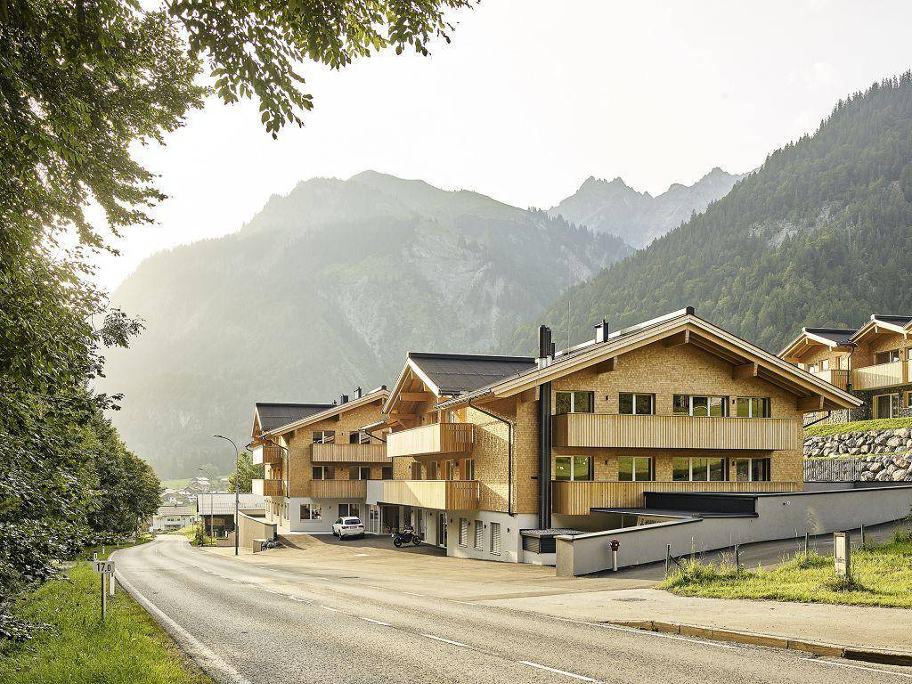 Moderne Chalets mit Holzbalkonen und Steinfassaden an einer Straße in einer Berglandschaft.