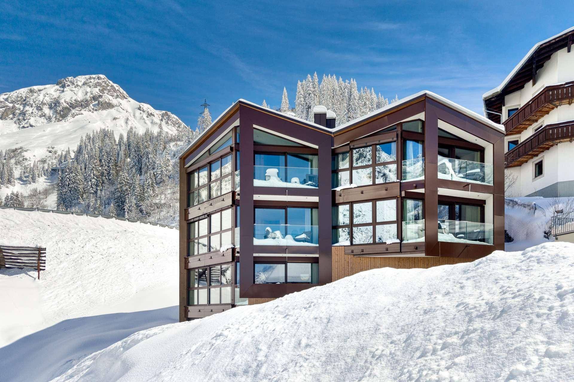 Modernes Gebäude mit großen Glasflächen und Balkonen in einer verschneiten Berglandschaft.