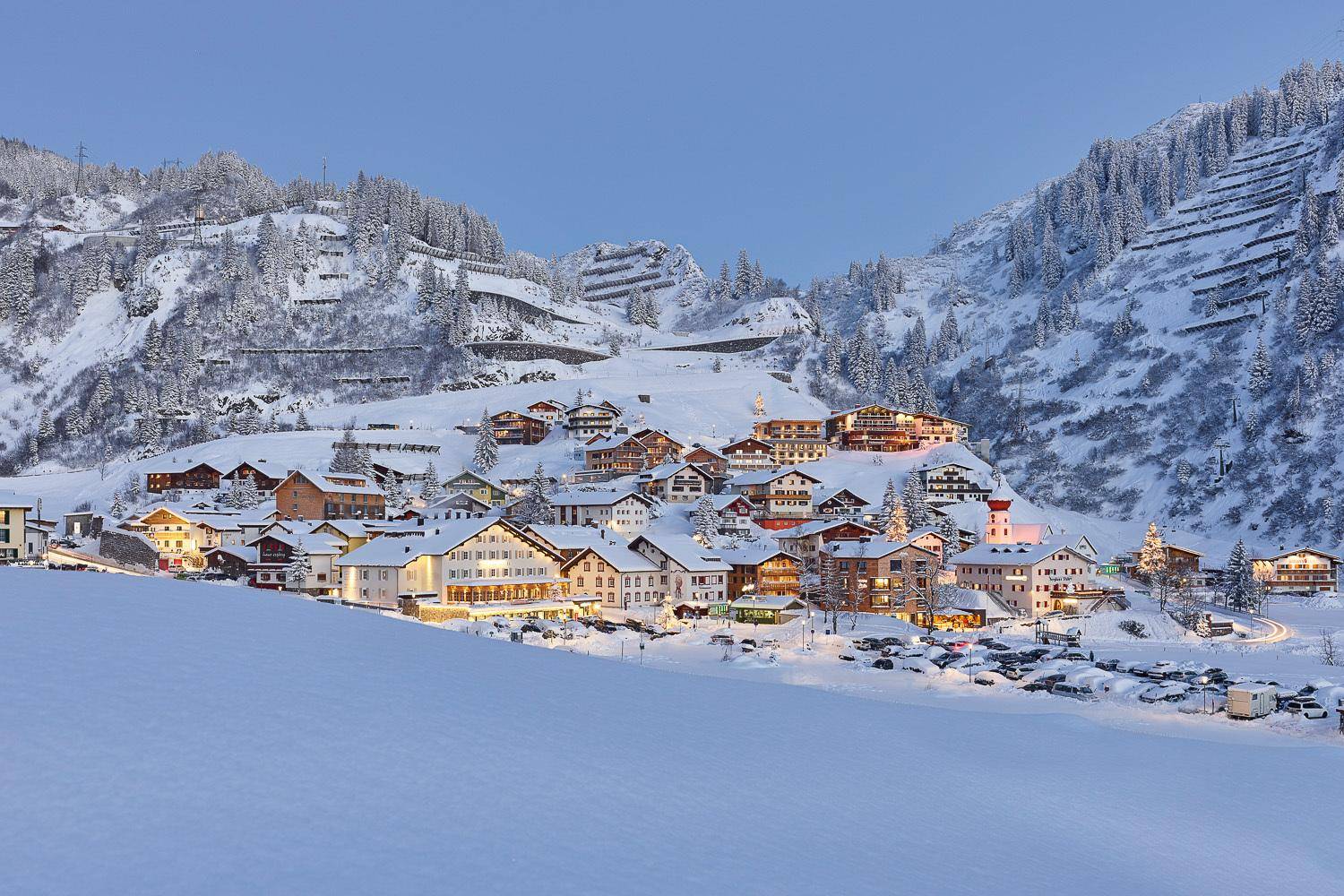 Malerisches Bergdorf in einer verschneiten Winterlandschaft mit beleuchteten Häusern bei Dämmerung.