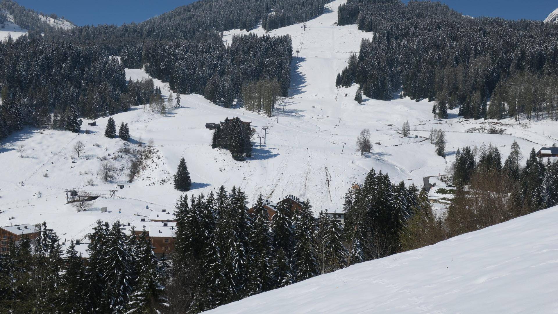 Winterliche Berglandschaft mit verschneiten Bäumen, Skipisten und einigen Gebäuden im Tal.