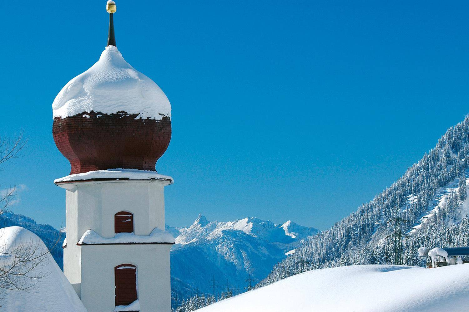 Eine schneebedeckte Kirchturmkuppel vor einer beeindruckenden Kulisse verschneiter Berge.