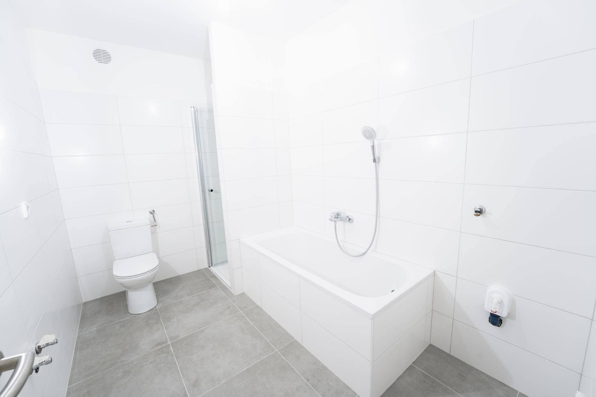 Helles, gefliestes Badezimmer mit Badewanne, Duschbereich und Toilette, unmöbliert.