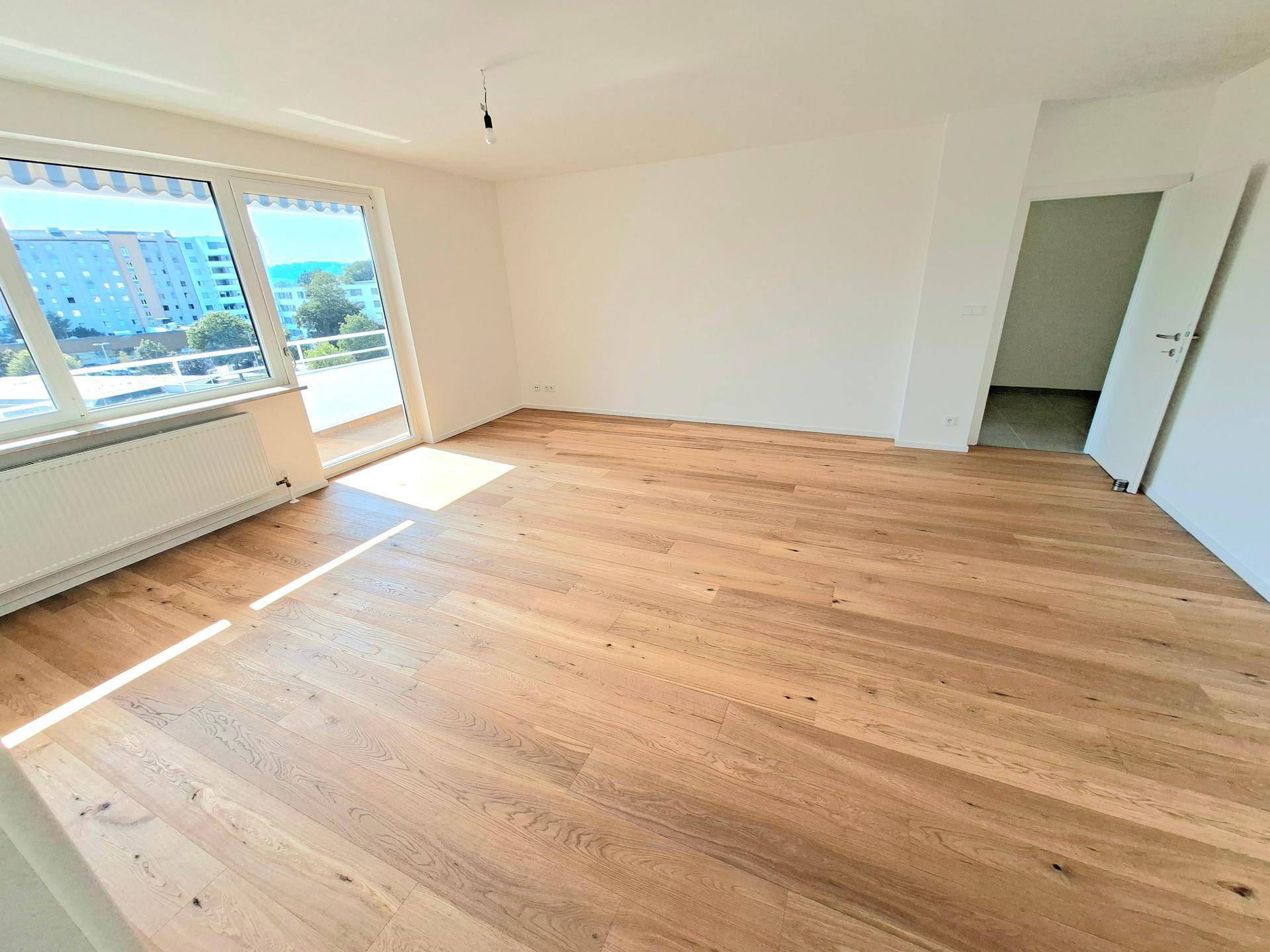 Geräumiger, leerer Wohn-Essbereich mit Holzboden und Zugang zu einem sonnigen Balkon.