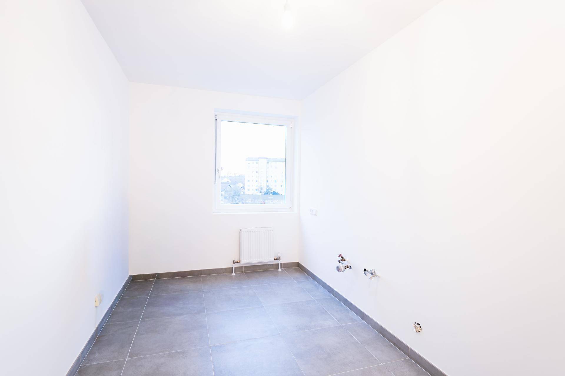 Leerer Küchenraum mit Fliesenboden, Fenster und Anschlüssen für eine zukünftige Einbauküche.