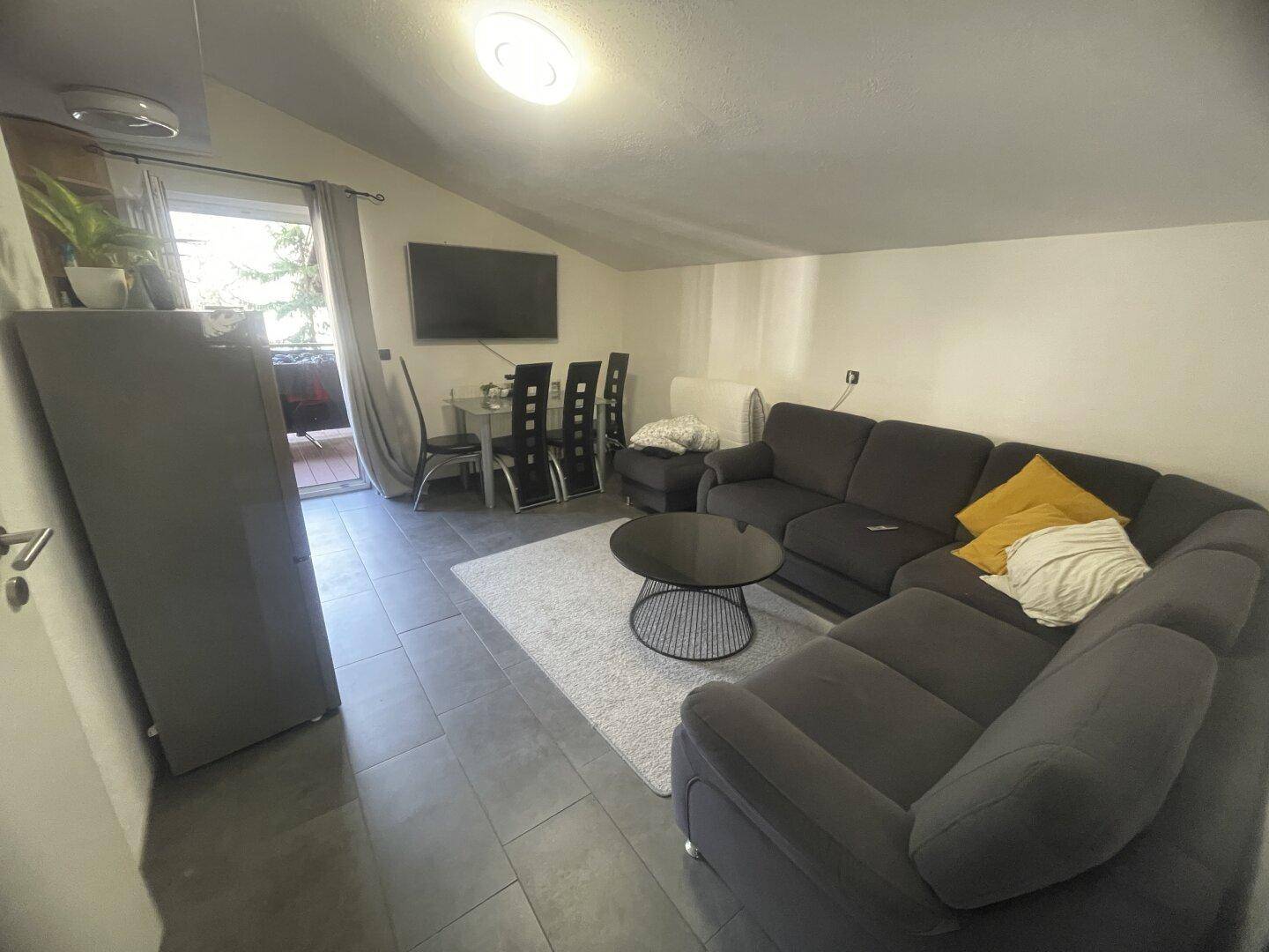 Gemütliches Wohnzimmer mit dunklem Ecksofa, Fliesenboden und Zugang zum Balkon.