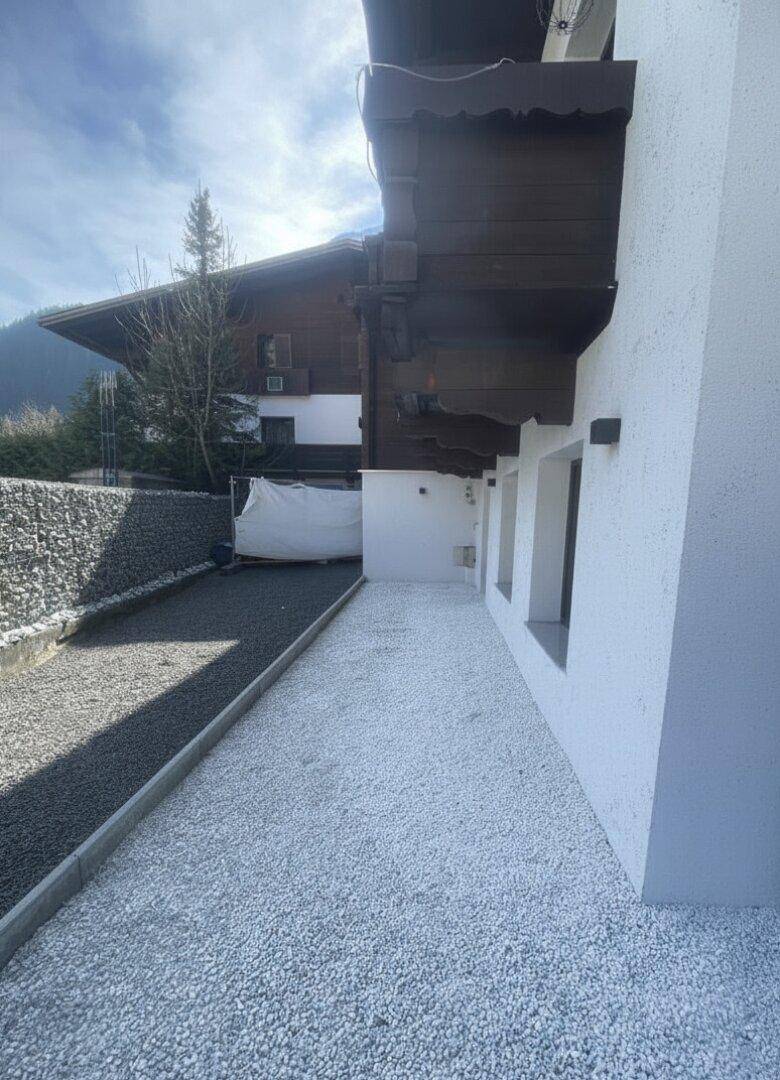 Kiesweg entlang der Hauswand mit Gabionenmauer und Blick auf die umliegende Bebauung.