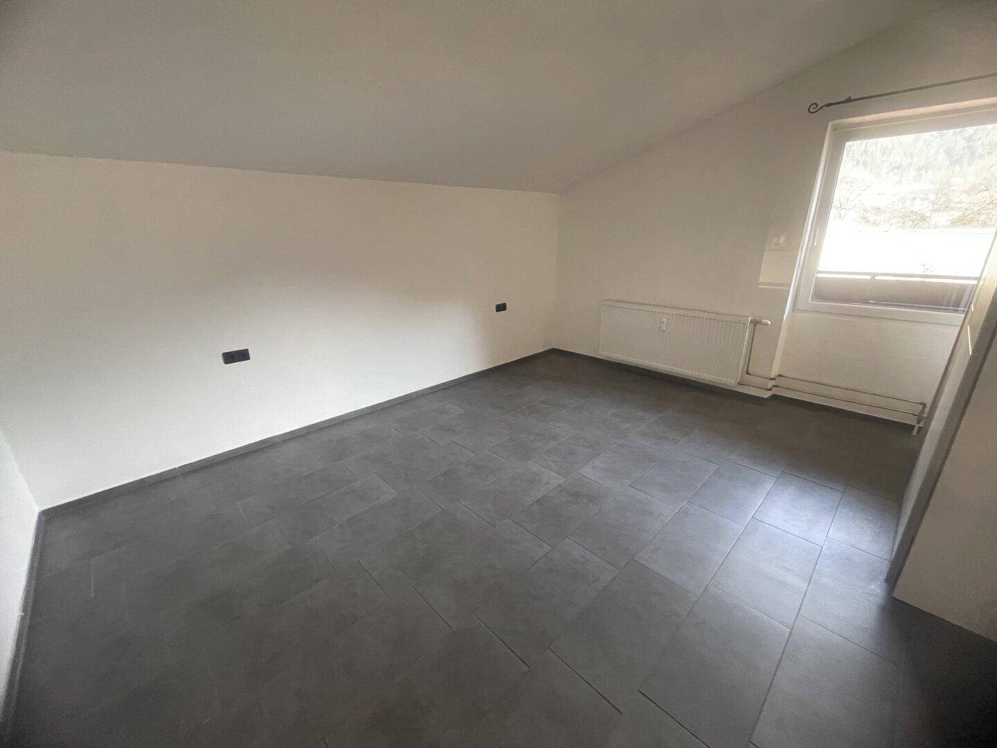 Leeres Zimmer mit Fliesenboden, Dachschräge und Fenster mit Blick nach draußen.