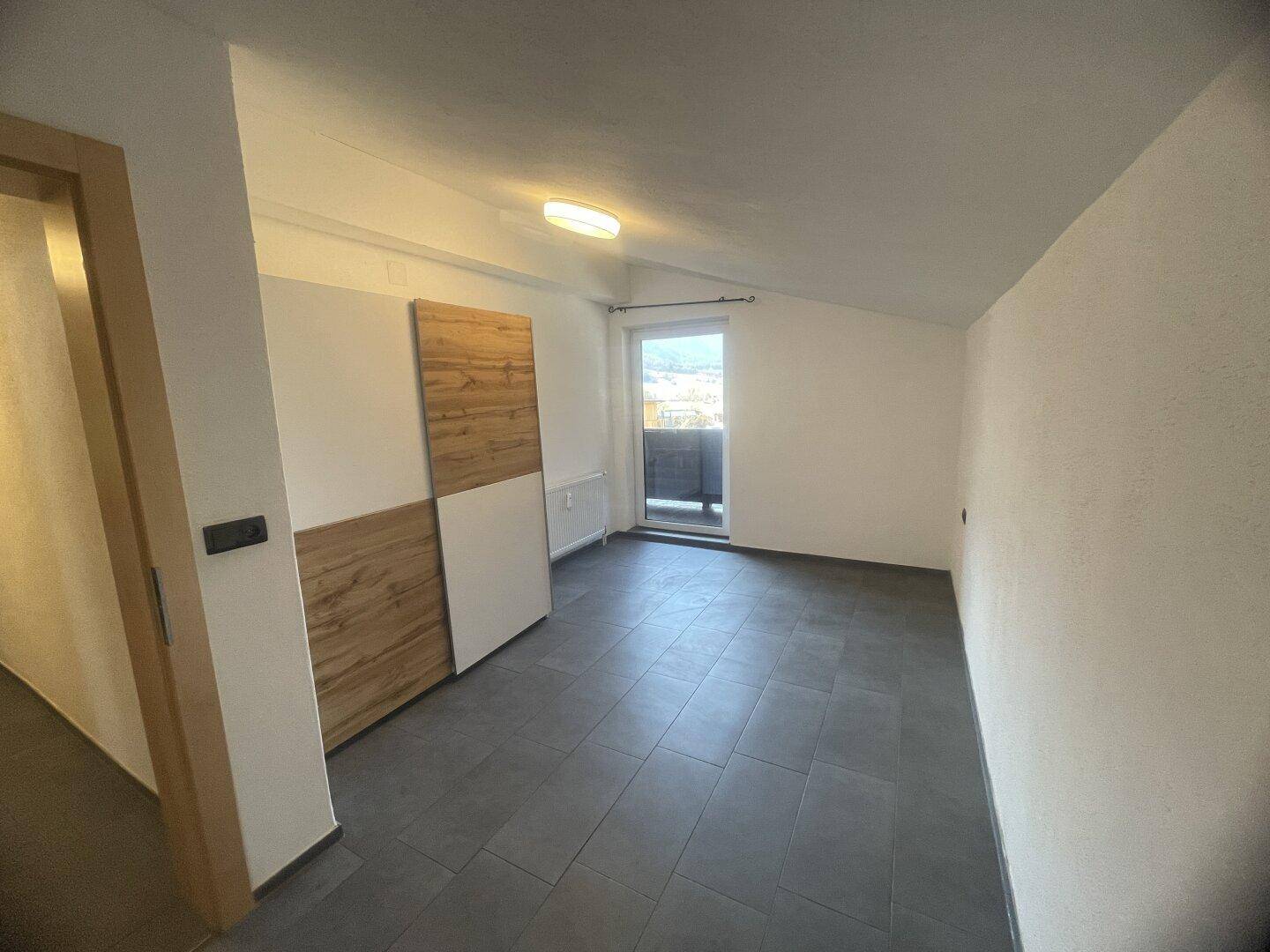 Helles Zimmer mit Fliesenboden, Dachschräge und Zugang zu einem kleinen Balkon.